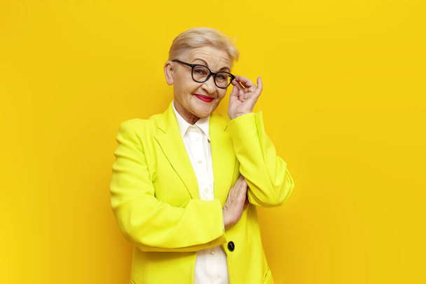 cheerful old business granny in blazer and glasses smiling on yellow isolated background, elderly businesswoman entrepreneur in formal wear - Photo, Image