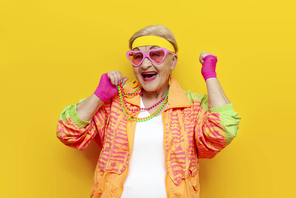 Sporcu hippi elbiseli komik yaşlı nine, sarı izole edilmiş arka planda başarı ve zafere seviniyor, genç kadın kıyafetleri içinde yaşlı kadın başarıyı kutluyor ve kazanıyor. - Fotoğraf, Görsel