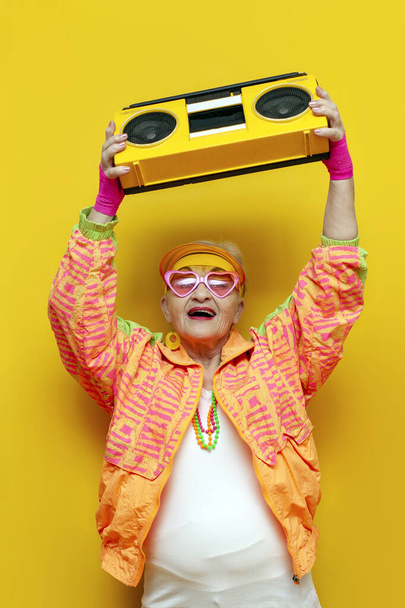 drôle vieille mamie folle avec magnétophone dans les vêtements de sport hipster écoute de la musique et chante sur fond jaune isolé, femme âgée dans des vêtements de jeunesse élégants avec lecteur de disques crie à la fête - Photo, image