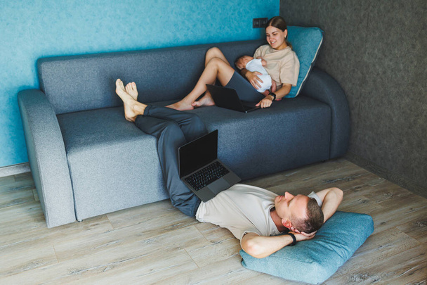 Γονείς με το μωρό κάθεται στον καναπέ στο σπίτι με φορητό υπολογιστή και μιλάμε στο διαδίκτυο. Μια νεαρή οικογένεια με ένα παιδί εργάζεται σε ένα φορητό υπολογιστή στο σπίτι, ενώ κάθεται στον καναπέ. - Φωτογραφία, εικόνα