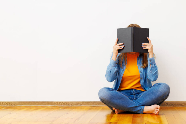 Μια νεαρή γυναίκα καλύπτει το πρόσωπό της με ένα βιβλίο καθώς κάθεται στο πάτωμα στον τοίχο.. - Φωτογραφία, εικόνα