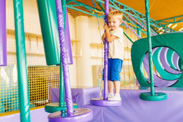 Милый маленький мальчик ползает и играет на красочной игровой площадке в закрытом парке развлечений - Фото, изображение