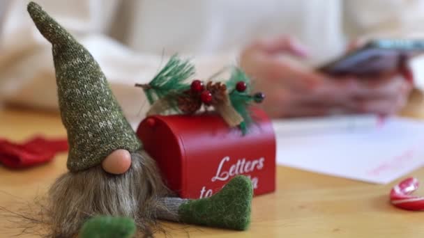 Um gnomo de brinquedo com uma caixa de correio vermelha para cartas do Papai Noel está na mesa, e uma adolescente sentada com um telefone em suas mãos em um fundo turvo, vista lateral close-up. Movimento lento com zoom - Filmagem, Vídeo