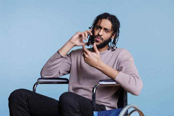 Hombre árabe confundido con impartición física que responde a la llamada del teléfono inteligente mientras está sentado en silla de ruedas y mirando a la cámara. Persona con discapacidad que habla en el retrato del teléfono móvil - Foto, Imagen