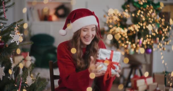 Захоплена молода жінка в капелюсі Санта грає з подарунковою коробкою в прикрашеному будинку під час Різдва - Кадри, відео