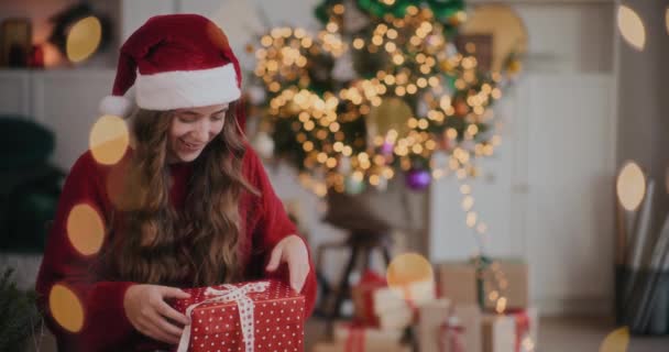 Веселая молодая женщина в шляпе Санты ловит рождественские подарки в украшенном доме - Кадры, видео