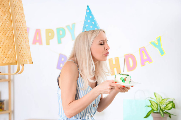 Mujer madura con pastel de cumpleaños haciendo deseo en la cocina - Foto, Imagen