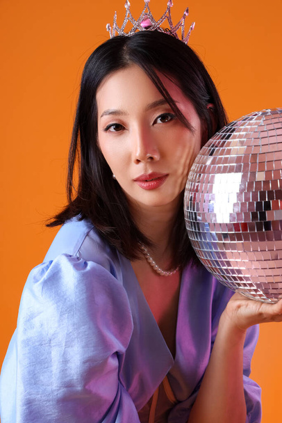 オレンジ色の背景にディスコボールを持つファッショナブルな美しいアジアの女性 - 写真・画像