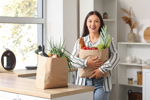 Jovem mulher asiática com sacos de compras cheios de comida fresca à mesa na cozinha - Foto, Imagem