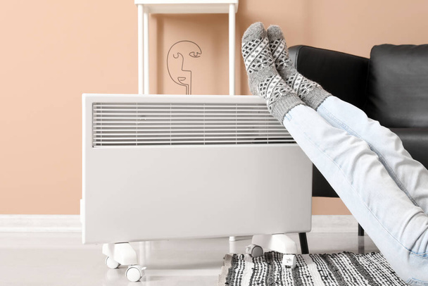 Νεαρή γυναίκα με ζεστές κάλτσες κοντά σε ηλεκτρική θερμάστρα στο σπίτι. Έννοια της εποχής θέρμανσης - Φωτογραφία, εικόνα