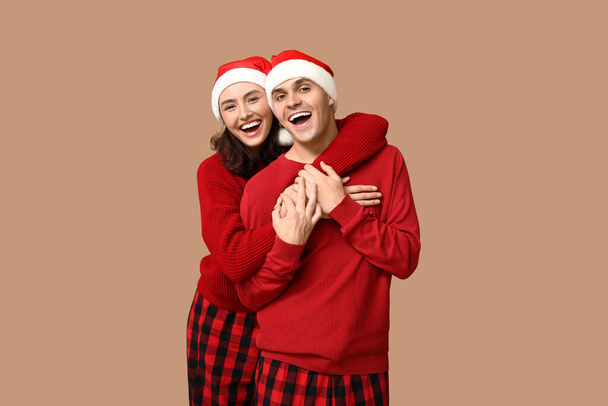 Happy young couple in Christmas pajamas and Santa hats hugging on beige background - Photo, Image