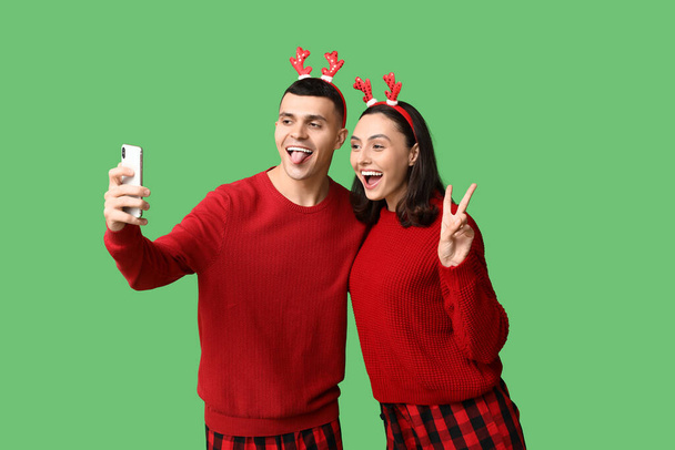 Happy young couple in Christmas pajamas taking selfie on green background - Photo, Image