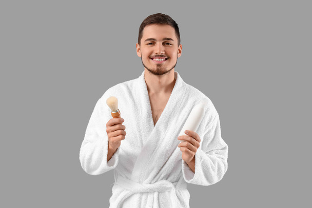 Handsome young man in bathrobe with shaving brush on grey background - Photo, Image
