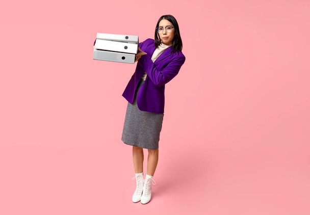 Портрет раздраженной азиатской бизнес-леди с офисными папками на розовом фоне - Фото, изображение