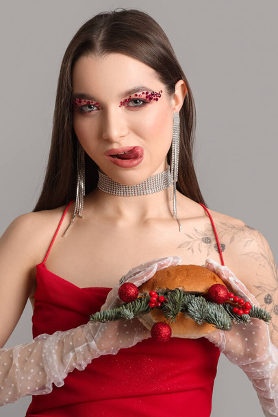 Beautiful young woman holding burger with Christmas balls and fir branches on grey background - Photo, Image