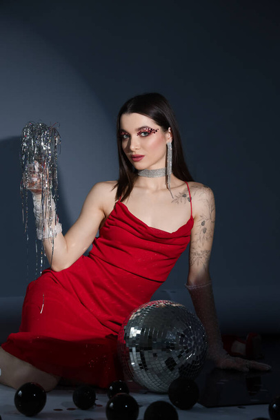 Young woman with disco ball and glass of Christmas tinsel sitting on grey background - Photo, Image