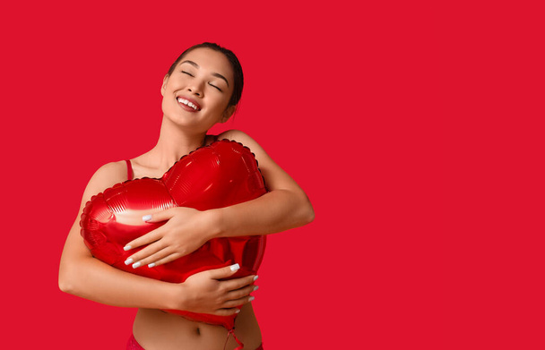 Όμορφη νεαρή ευτυχισμένη Ασιάτισσα γυναίκα σε σέξι εσώρουχα με μπαλόνι σε σχήμα καρδιάς σε κόκκινο φόντο. Γιορτή του Αγίου Βαλεντίνου - Φωτογραφία, εικόνα