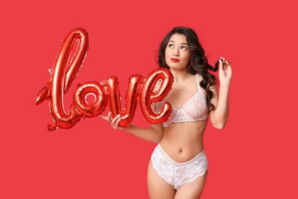 Όμορφη νεαρή στοχαστική Ασιάτισσα γυναίκα με μπαλόνι σε σχήμα λέξης ΑΓΑΠΗ φορώντας σέξι εσώρουχα σε κόκκινο φόντο - Φωτογραφία, εικόνα
