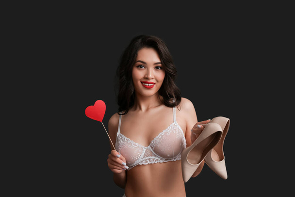 Όμορφη νεαρή ευτυχισμένη Ασιάτισσα γυναίκα σε σέξι εσώρουχα με χάρτινη καρδιά και κομψά ψηλά τακούνια σε μαύρο φόντο - Φωτογραφία, εικόνα