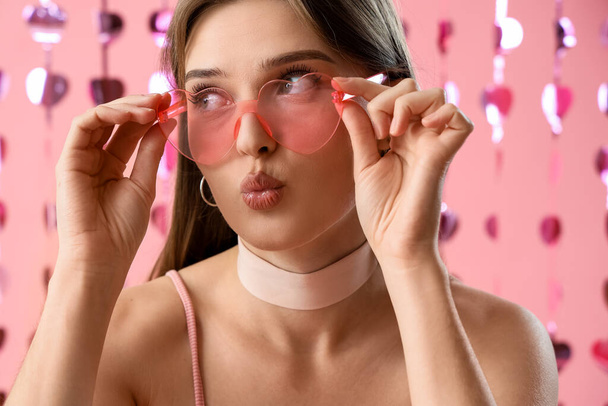 Young woman in heart-shaped sunglasses blowing kiss on pink background, closeup. Valentine's Day celebration - Photo, Image