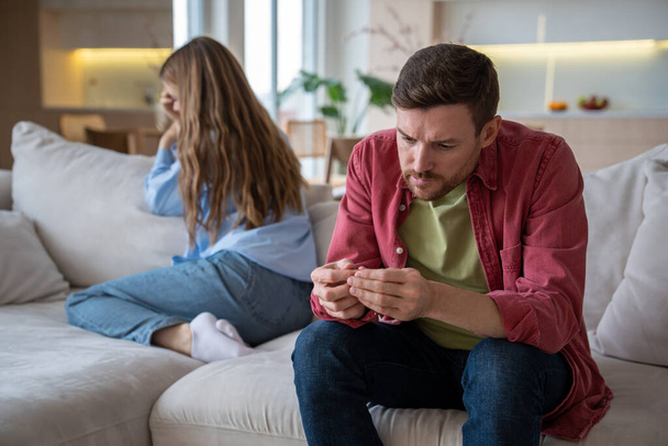 Relationships crisis, misunderstanding, toxic partners. Spouses ignoring each other after quarrel manipulating silence sitting on sofa at home. Nervous man examines nails on hands woman turning away. - Photo, Image