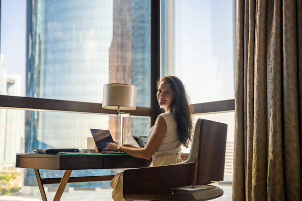 Стильная деловая женщина фрилансер, работающая с ноутбуком, сидит на стуле, панорамный вид в окне с городским фоном. Фото с низким ключом. Высокое качество фото - Фото, изображение