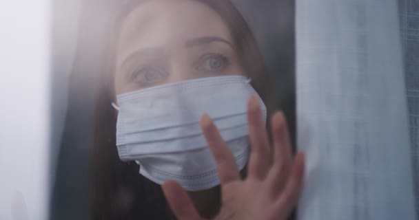 Face mask, thinking and woman by window sad in home for reflection, remember and wonder. Quarantine, healthcare and face of person by glass with protection for pandemic, sickness and epidemic safety. - Footage, Video
