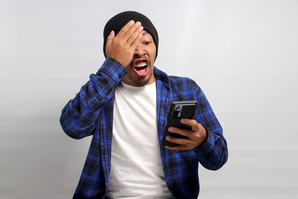 Shocked Asian man, dressed in a beanie hat and casual shirt, covers his eyes in a comical and surprised reaction while looking at his phone, clearly taken aback by unexpected news, white background - Photo, Image