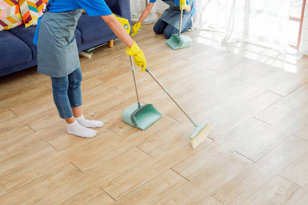 Wizerunek plonu młodej profesjonalnej sprzątaczki pracującej w domu. Gospodyni zamiata miotły na drewnianej podłodze i sprząta pod kanapą. Koncepcja czyszczenia i prac domowych - Zdjęcie, obraz