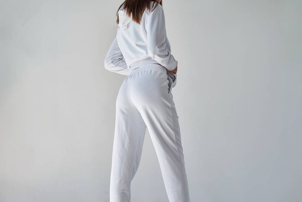 この現代的なホワイトアンサンブルでカジュアルスタイルの究極を体験してください. スポーツウェアモックアップ. 白いズボンの女の子。 白いミニマリストのスウェットパンツを着ている若い女性のバックビュー. - 写真・画像