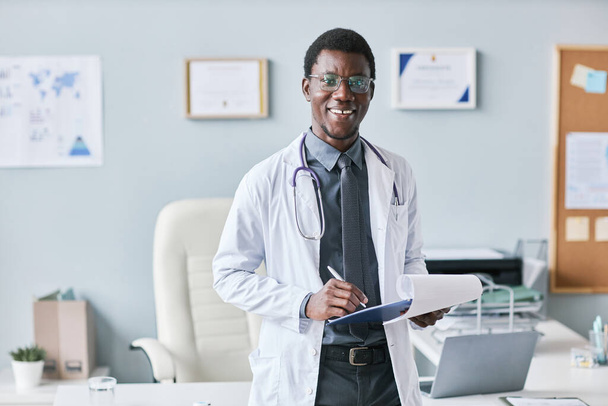 Retrato de la cintura hacia arriba del joven médico afroamericano de pie junto al escritorio en la clínica y sujetando el portapapeles sonriendo a la cámara - Foto, Imagen