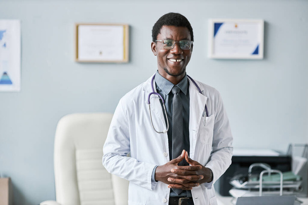 Taille omhoog portret van jonge Afro-Amerikaanse arts in het kantoor staan en glimlachen op camera, kopieer ruimte - Foto, afbeelding