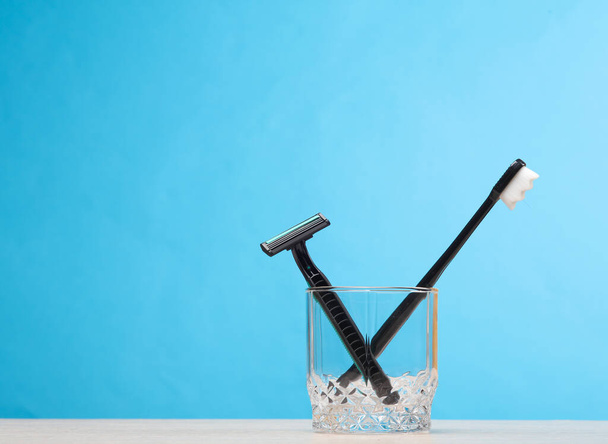 Lâmina descartável e escova de dentes em um copo na mesa, fundo azul. rotina diária, atendimento odontológico e barbear - Foto, Imagem