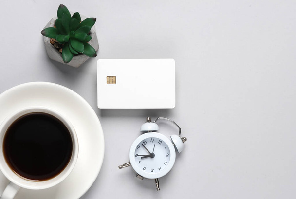 Weiße leere Bankkarte mit Chip, Tasse Kaffee, Pflanze und Wecker auf grauem Hintergrund. Flaches Laien-Geschäftskonzept. Ansicht von oben - Foto, Bild