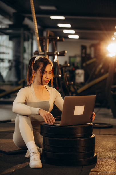 A woman communicates with a trainer through a laptop and works out in the gym - Photo, Image