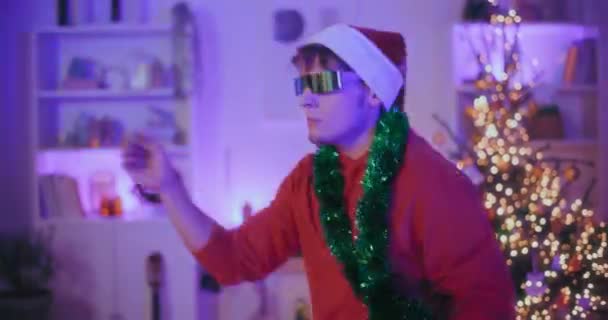 Aufgeregter junger Mann mit Nikolausmütze und Sonnenbrille tanzt zu Weihnachten vor beleuchtetem Haus - Filmmaterial, Video