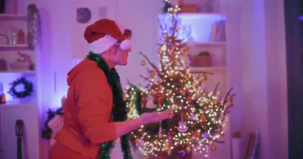 Joven en sombrero de Santa y gafas de sol con oropel verde en el hombro bailando en casa iluminada durante la Navidad - Metraje, vídeo