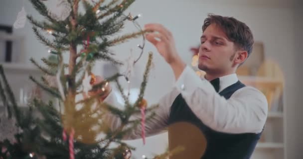 Ben vestito giovane uomo mettendo le luci delle fate sull'albero di Natale a casa - Filmati, video