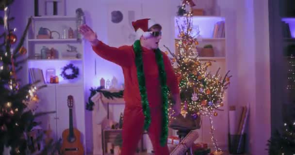 Joven excitado en Santa sombrero y gafas de sol bailando en casa iluminada durante la Navidad - Metraje, vídeo