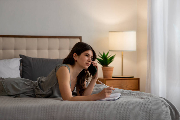 Mooi meisje liggend op bed met dagboek en pratend aan de telefoon. Jonge vrouw met notitieboekje en pen. Hoge kwaliteit foto - Foto, afbeelding