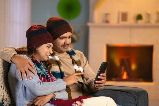Счастливые пары с зимней одеждой, пользуясь мобильным телефоном на диване дома - концепция технологии, связи и привязанности. - Фото, изображение
