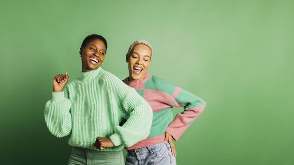 Dos jóvenes mujeres alegres vistiendo ropa verde y bailando en un estudio con un fondo verde. - Foto, imagen