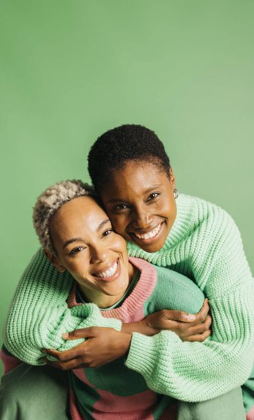 Two young playful women wearing green clothes embracing in a studio with a green background. - Photo, Image