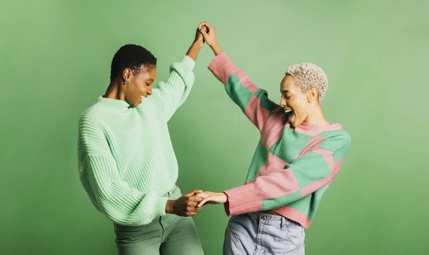 Two young cheerful women wearing green clothes and dancing in a studio with a green background. - Photo, Image