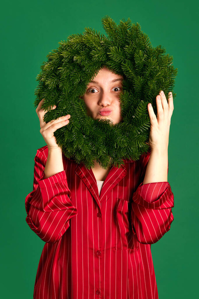 Portré gyönyörű fiatal lány piros pizsamában mosolyog és grimaszok tartó szilveszteri koszorú zöld stúdió háttér. A boldog új év koncepciója, boldog karácsony, ünnepi hangulat. - Fotó, kép