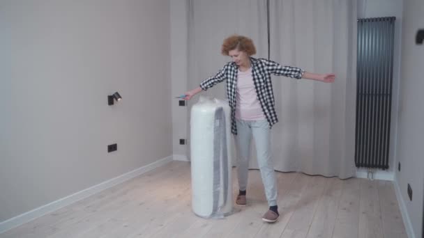 Housewife opens a newly purchased latex mattress from a vacuum roll without air. Mature woman unpacking mattress in new empty apartment without furniture while relocating to new home. Move-in.  - Footage, Video