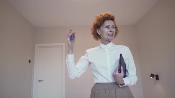 Az idősebb női ingatlanügynöknél van az új lakás kulcsa. Érett nő Ingatlan bróker ad kulcsot a házhoz, miközben áll a háttérben az üres, kis hangulatos lakás kiadó. Ingatlanügyletek.  - Felvétel, videó