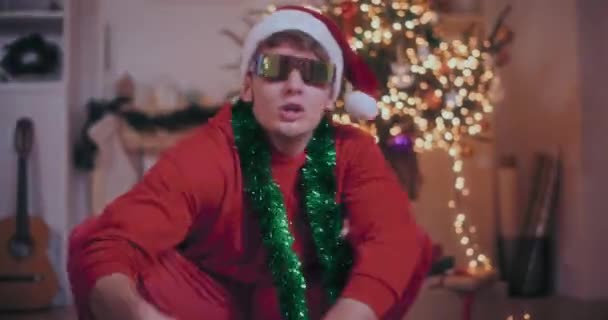 Nadšený mladý muž se zeleným tinsel kolem krku zpěv, zatímco dřepí v osvětlené domů během Vánoc - Záběry, video