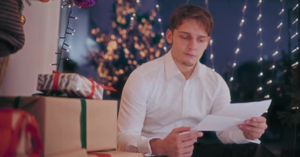 Resmi kıyafetli genç adam Noel hediyesini akşamları aydınlık evde okuduktan sonra açıyor. - Video, Çekim