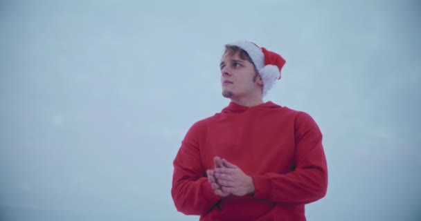 Вид з низьким кутом зору вдумливого молодого чоловіка в червоній куртці з капюшоном і капелюхом Санта, дивлячись далеко, стоячи проти неба під час Різдва - Кадри, відео